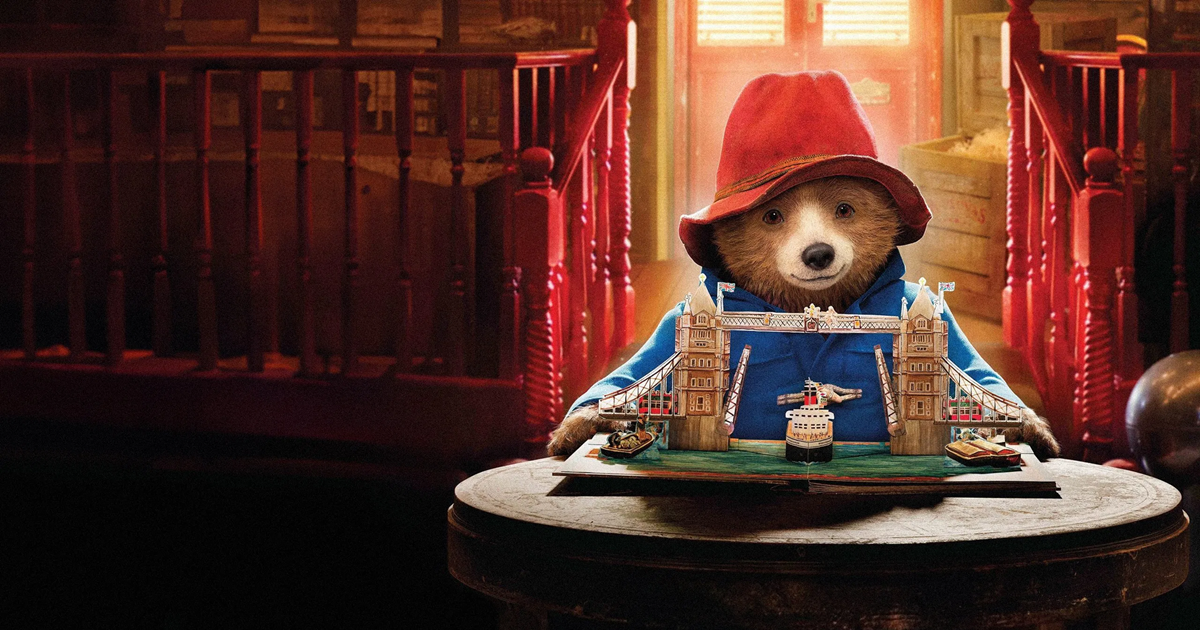 تصویری از پدینگتون، خرسی کوچک با کلاه قرمز و کت آبی، در حال لبخند زدن و ماجراجویی در شهر