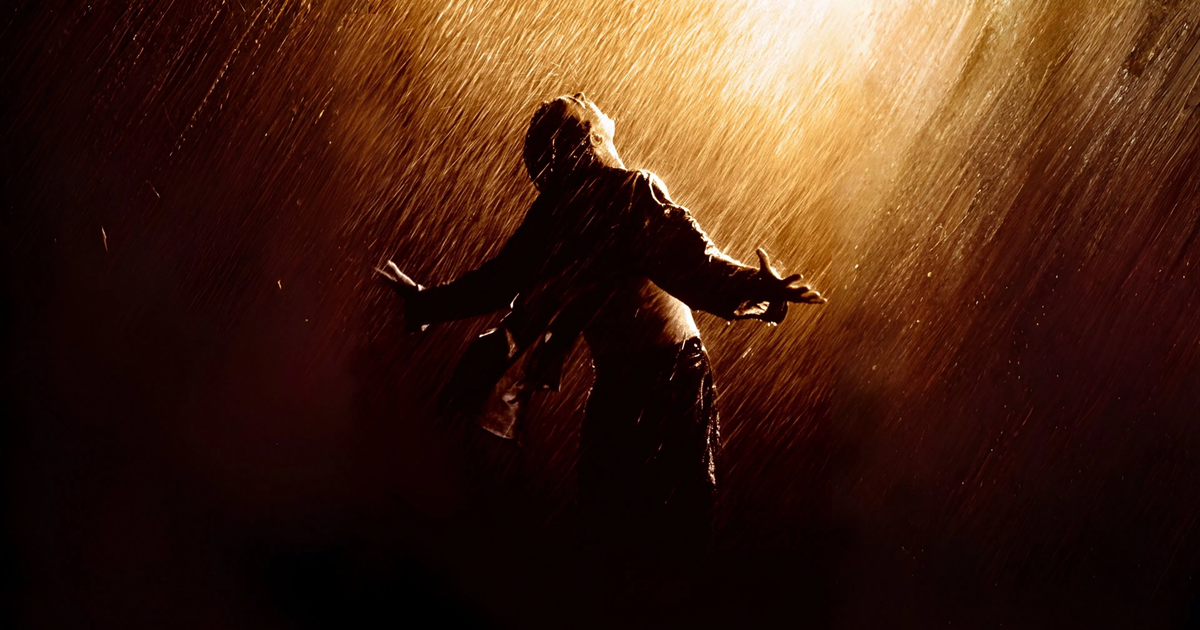 تصویری از اندی دوفرین، مردی با لباس زندان، ایستاده در زیر باران شدید با دستان باز به سمت آسمان